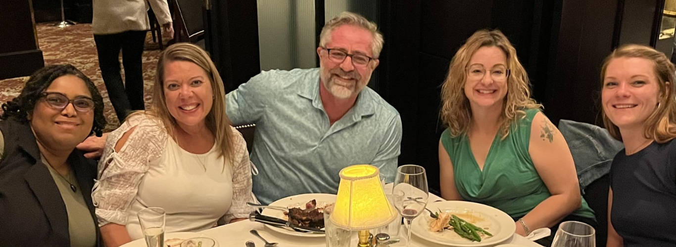 Five smiling adults sit together at a restaurant table with plates of food and drinks in front of them, posing for a group photo in a warmly lit dining room.
