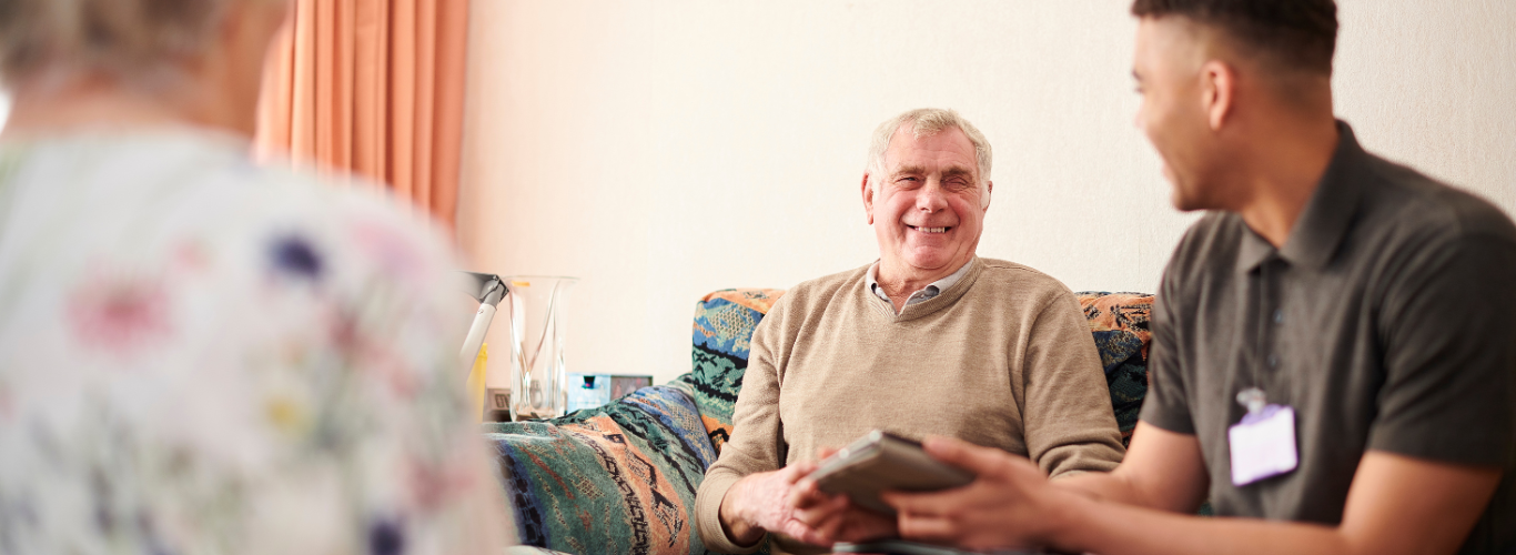 A person wearing a polo shirt holds a tablet while sitting on a couch next to another person who is smiling.